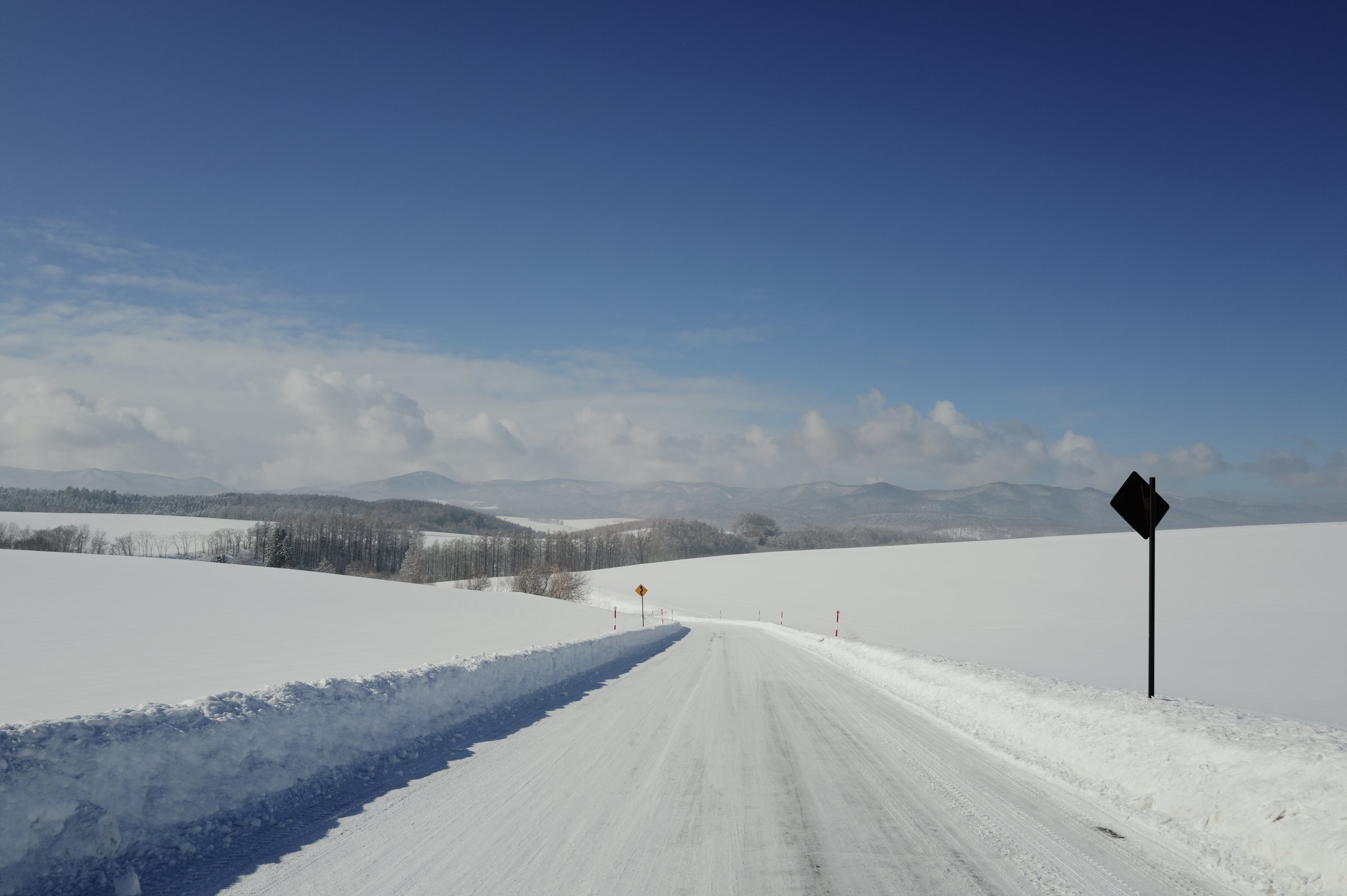 雪の丘のあいだを延びる道と、遠くの山並み