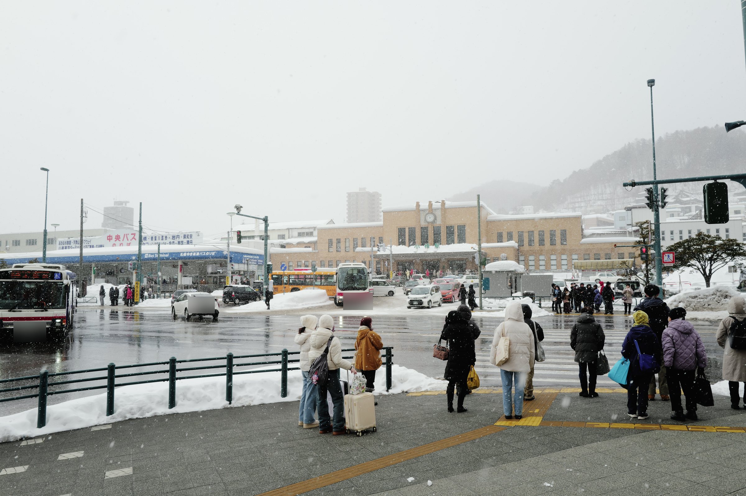 雪の降る駅前で信号を待つ人たちと、その向こうのバスと駅舎