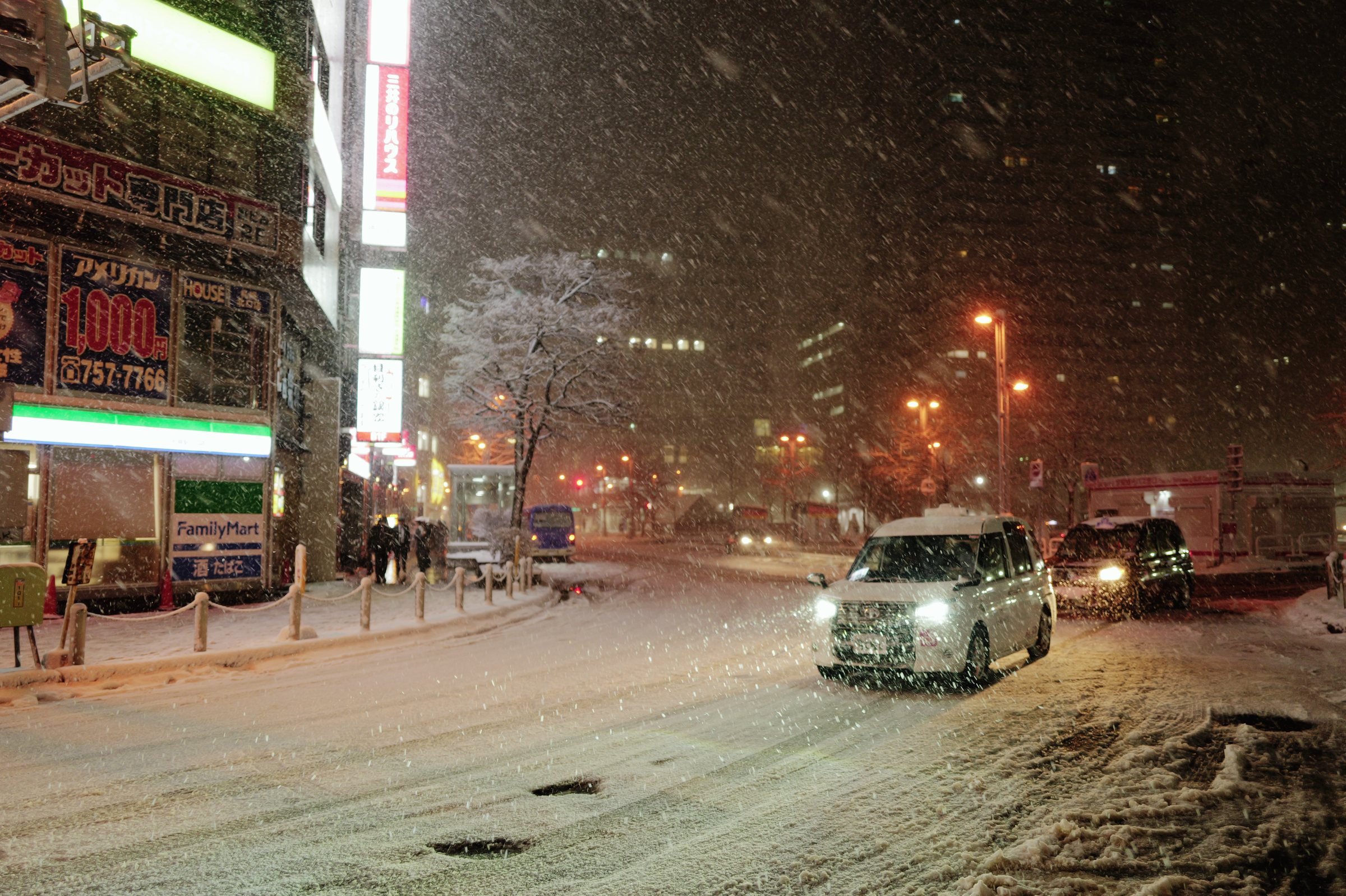 雪の降る夜、ライトをつけて駅前の広場を進む車