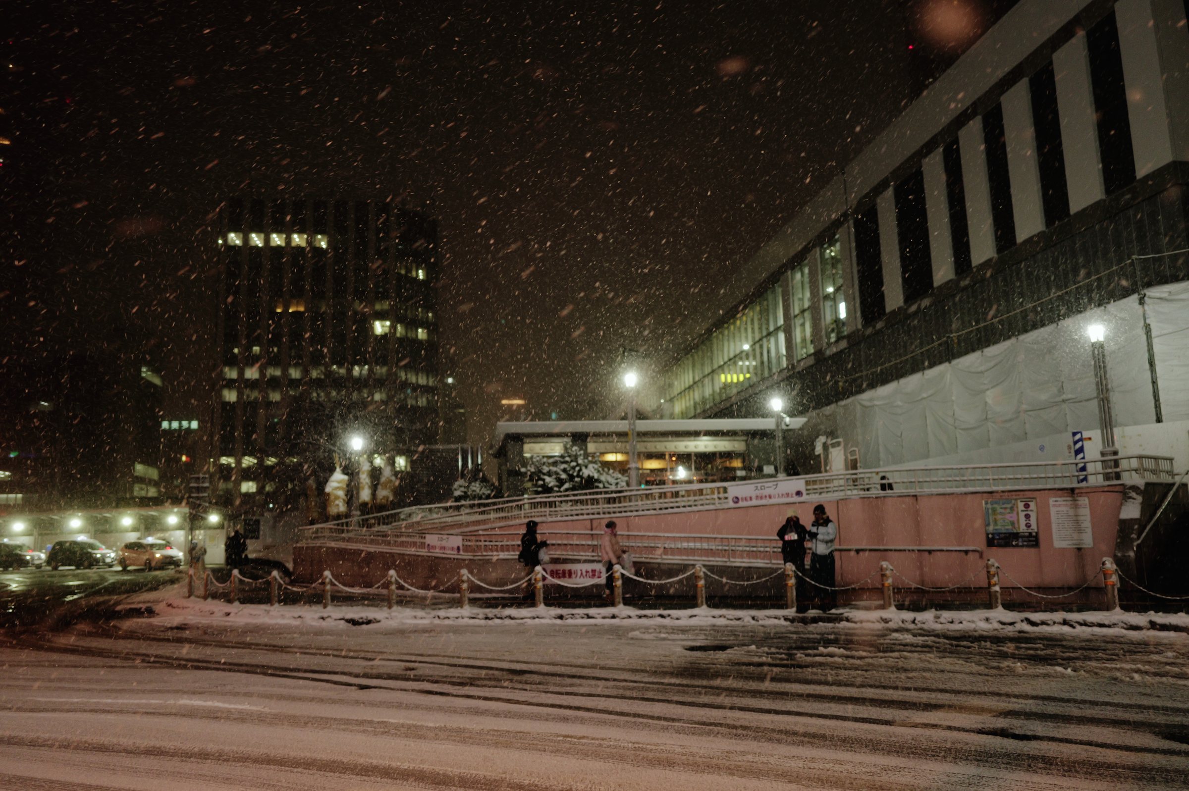 雪の降る夜の駅前広場と、灯りのともる駅の建物