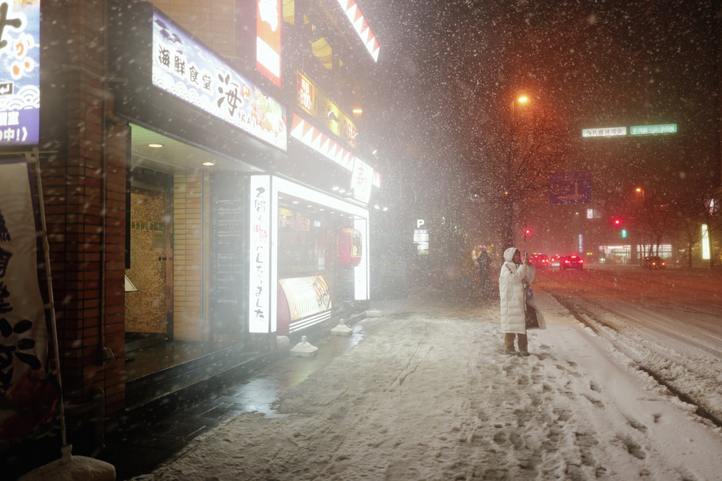 雪の降る夜の歩道、明るい店の前に立つ人と、橙色に染まる雪
