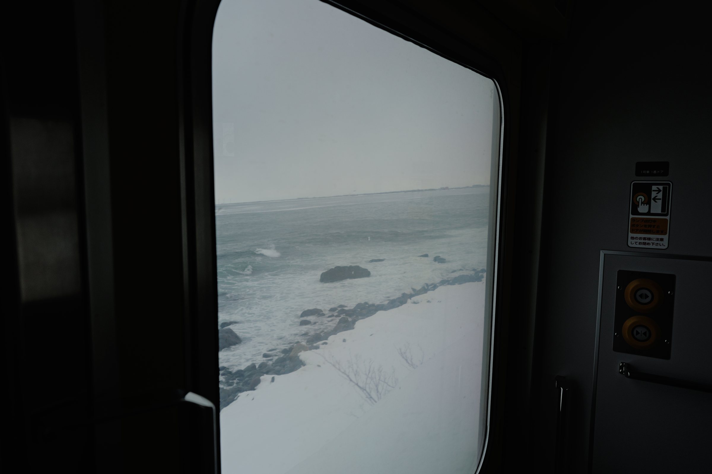 列車の窓の向こうの、雪をかぶった岩と灰色の海