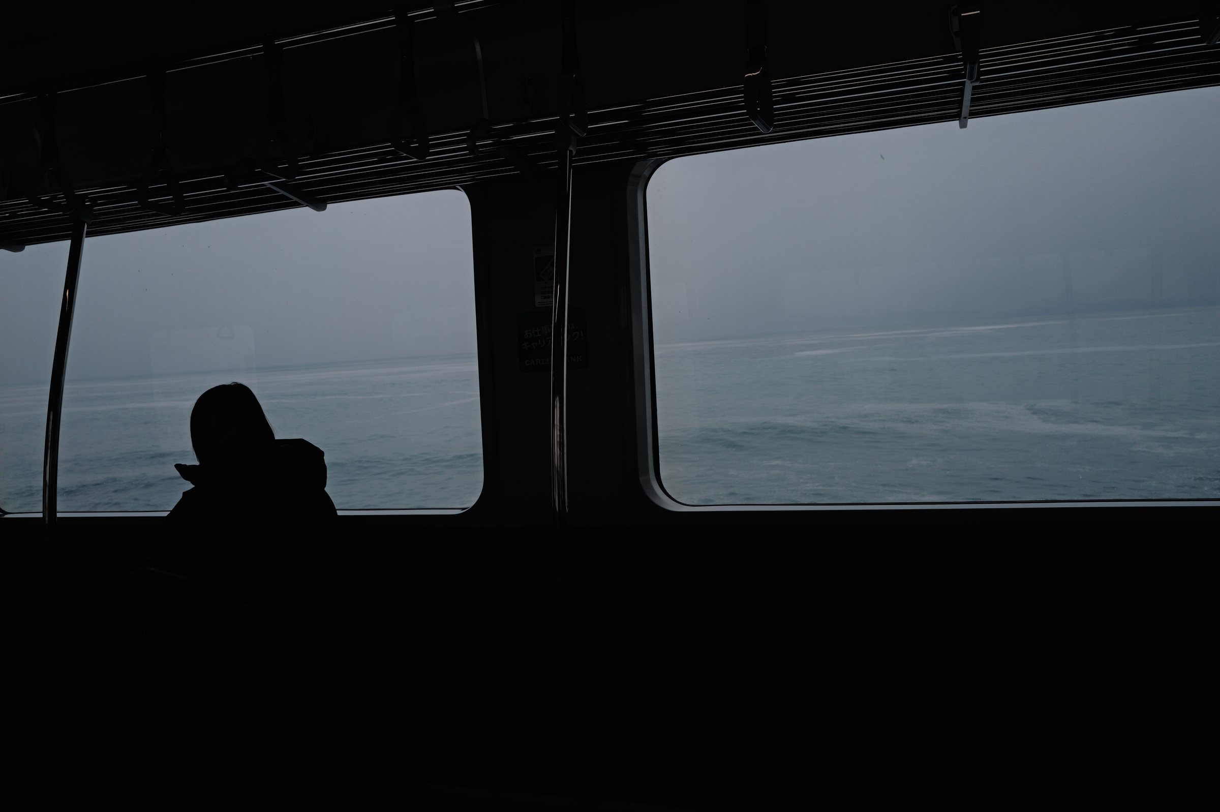 暗い車内で影になって座る人と、窓の外の灰色の海