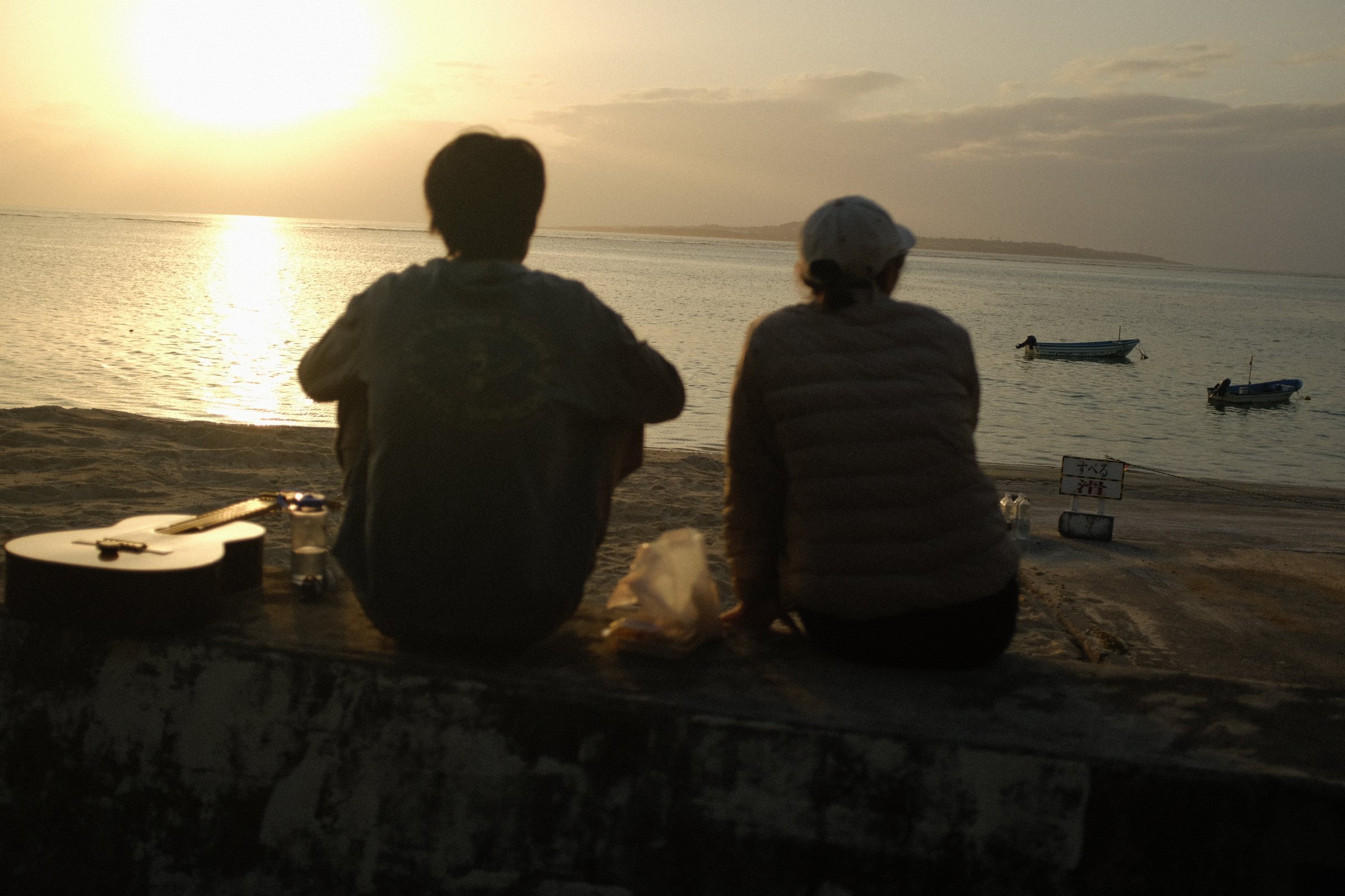 夕陽を前に、堤防に並んで座る二人の背中と、脇に置かれたギター