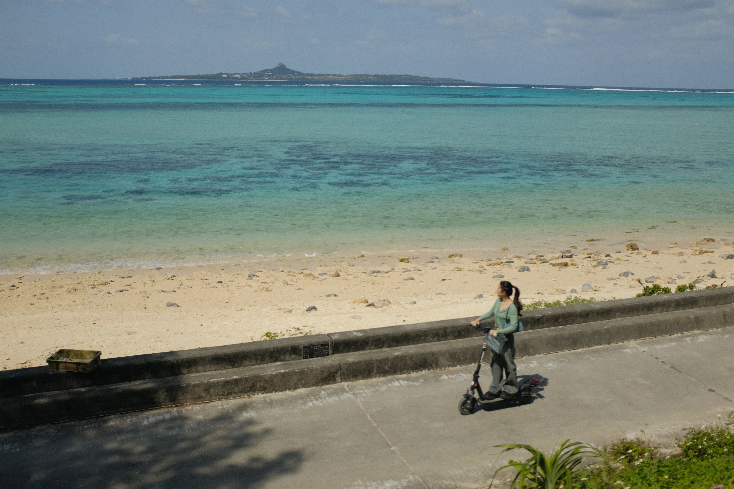 伊江島の見える青い海と、浜沿いの道を行くキックボードの人
