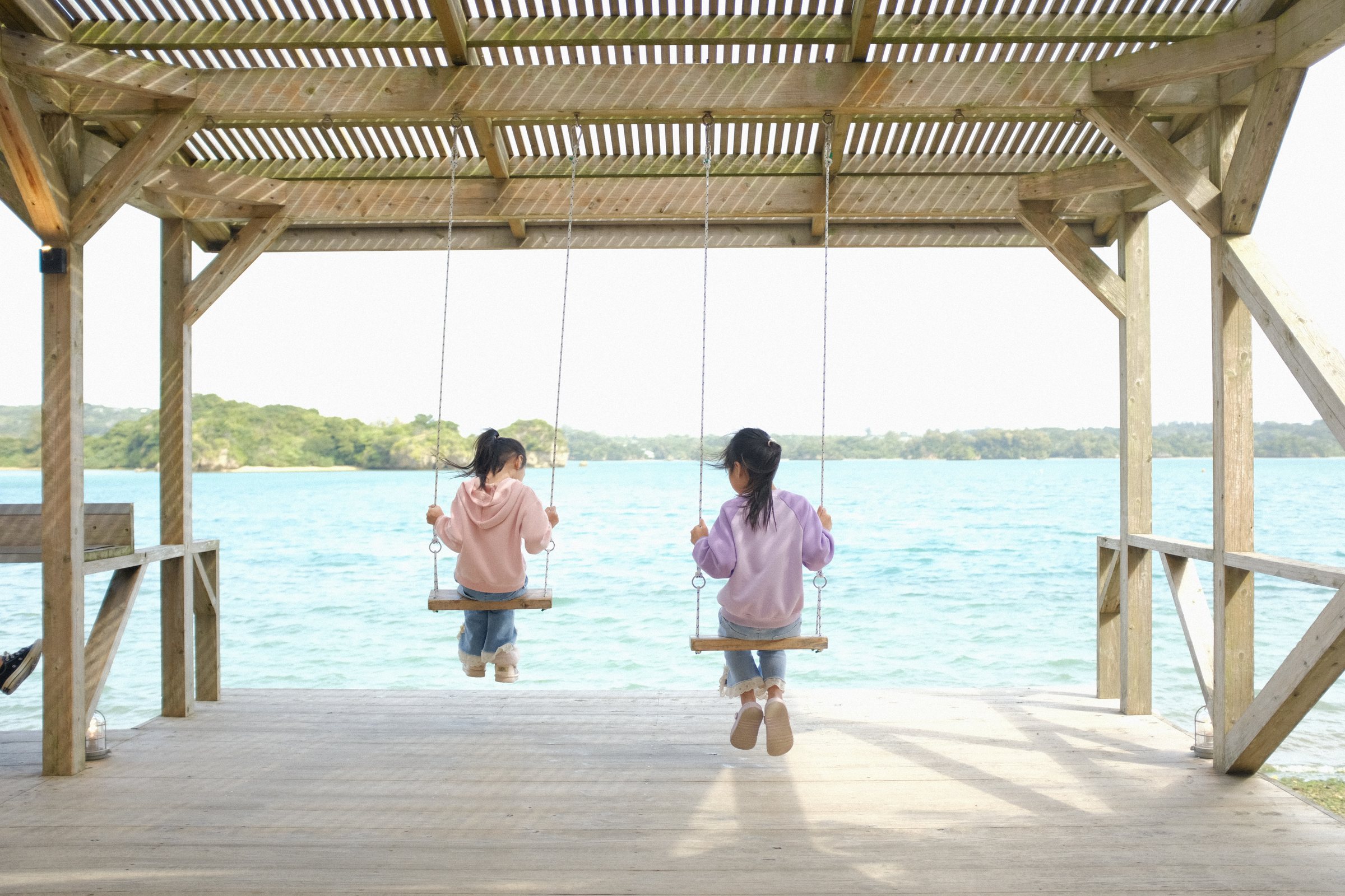 海に面した東屋のブランコで揺れる、二人の子どもの後ろ姿