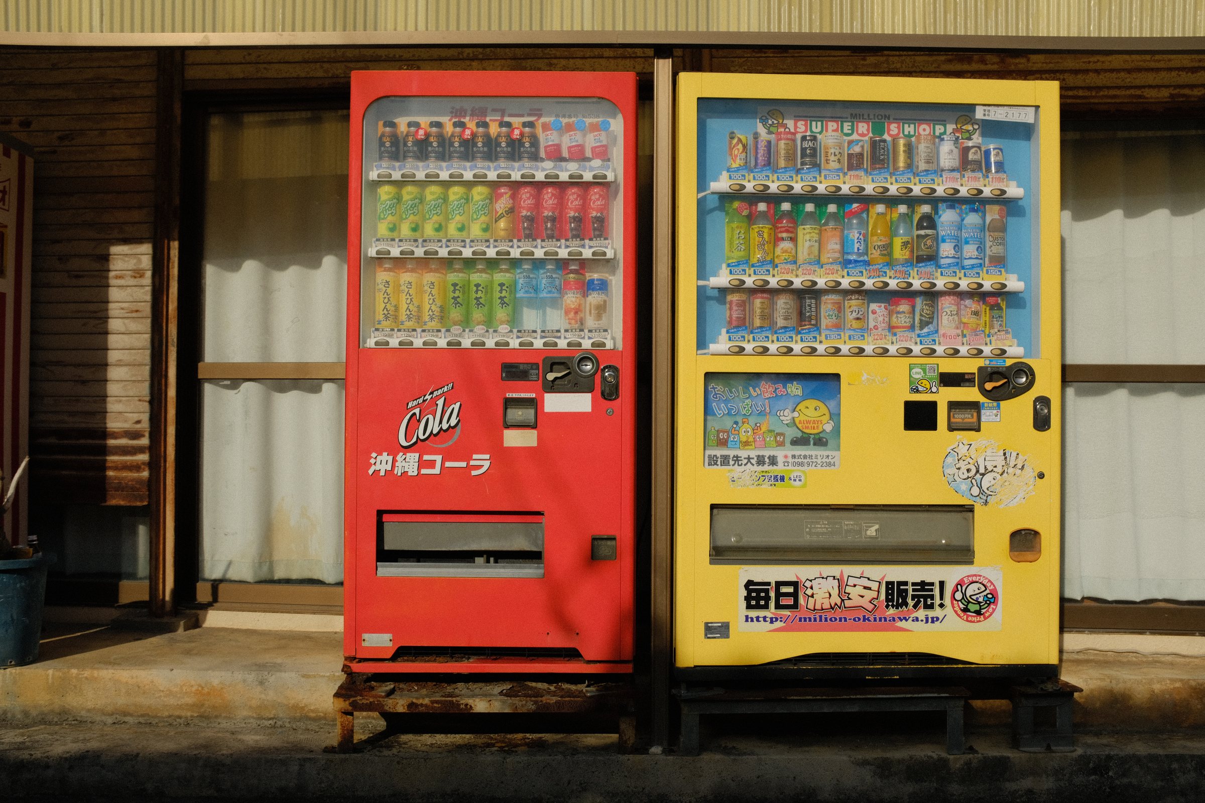 軒下に並ぶ赤い「沖縄コーラ」の自販機と黄色い自販機