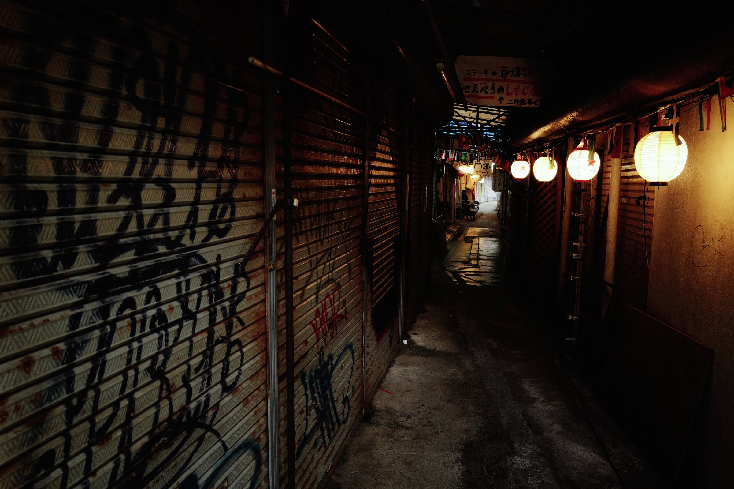 落書きのあるシャッターの路地と、奥に並ぶ提灯