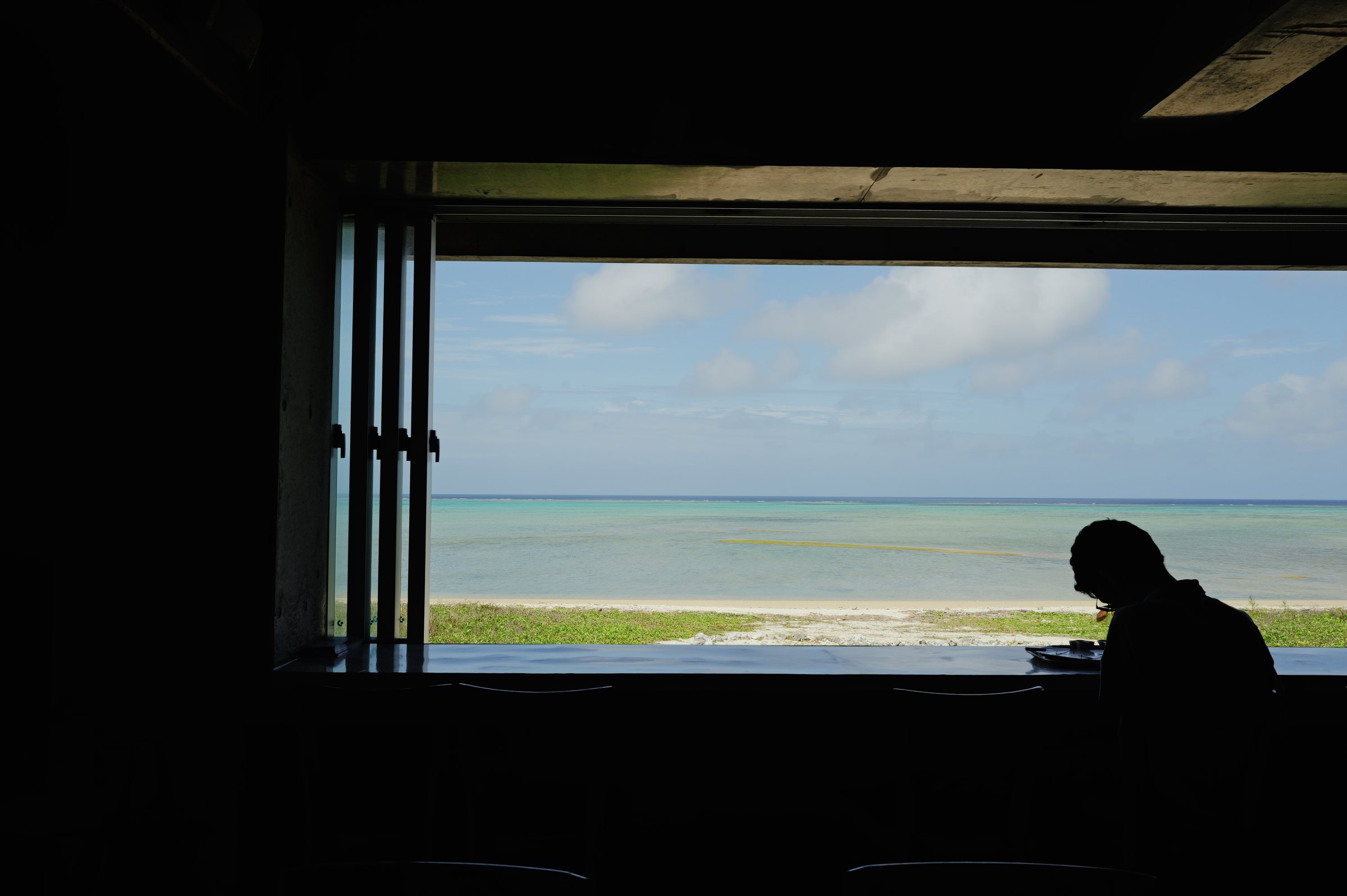 暗い店内の窓辺に座る人の影と、窓の外の白い浜と海