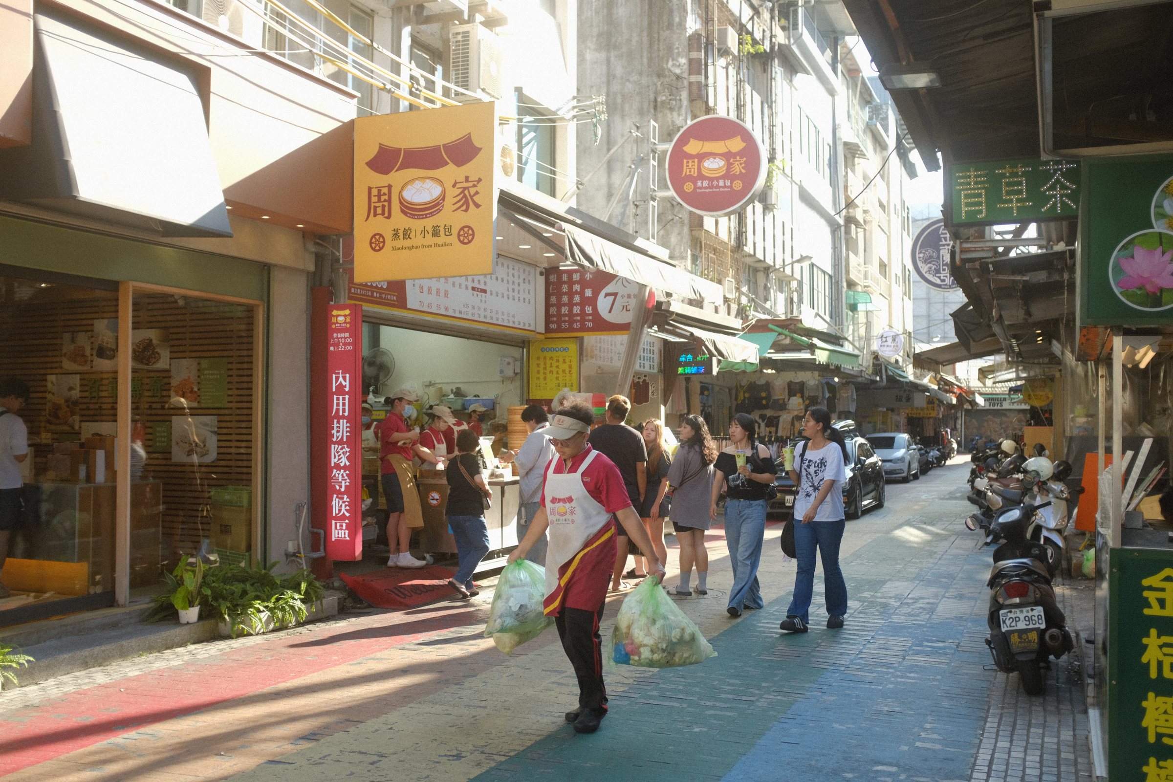 日の当たる商店街を、買い物袋を提げて歩く人たち