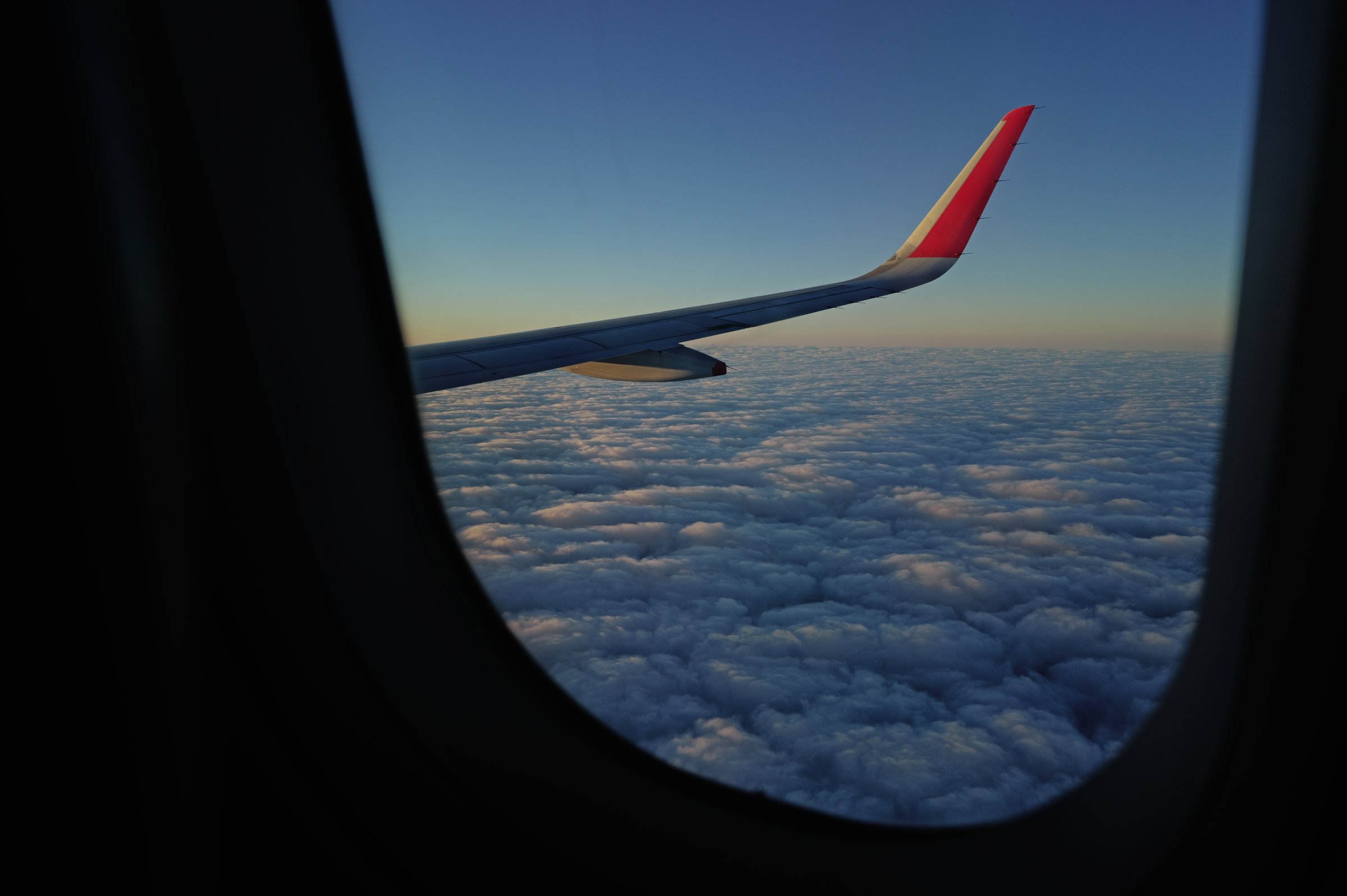 飛行機の丸い窓の向こうに、夕方の雲と赤い翼の先