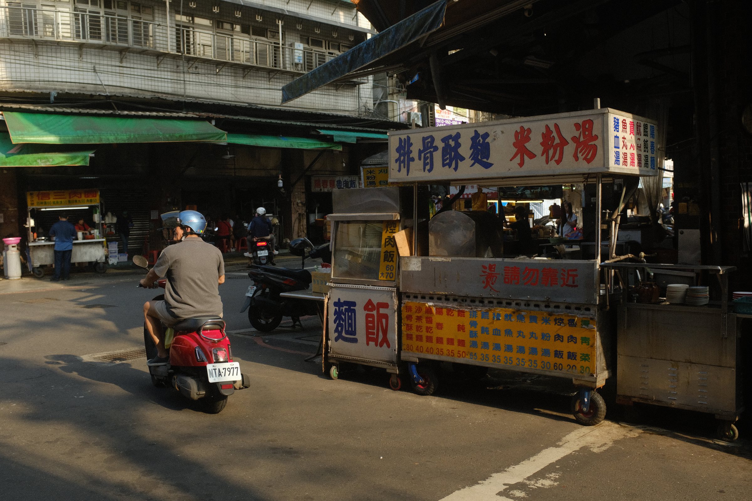 西日の差す通りで、麺と米粉湯の屋台の前をバイクが通り過ぎていく