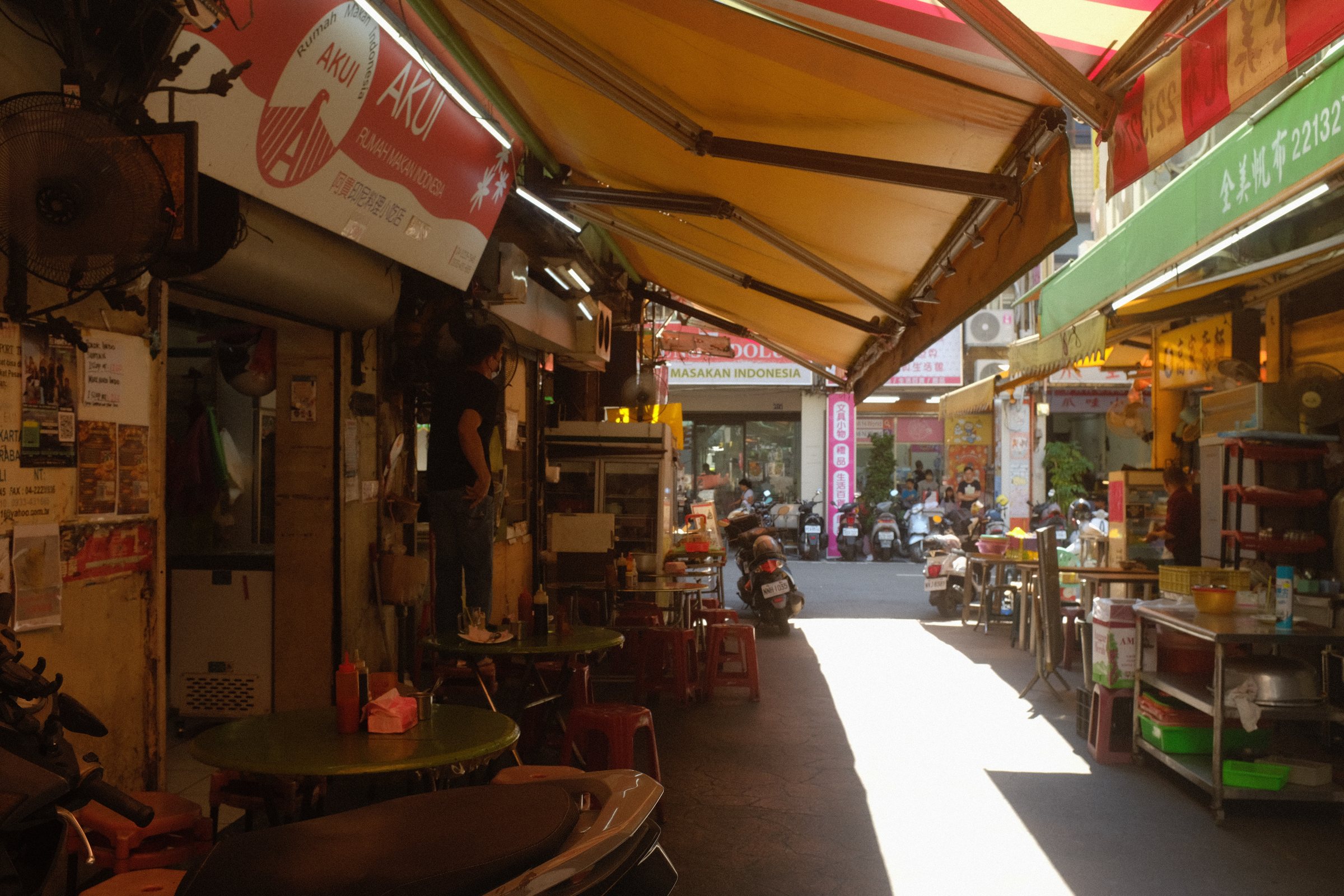 オレンジ色の日除けの下、光の差し込む路地の店先