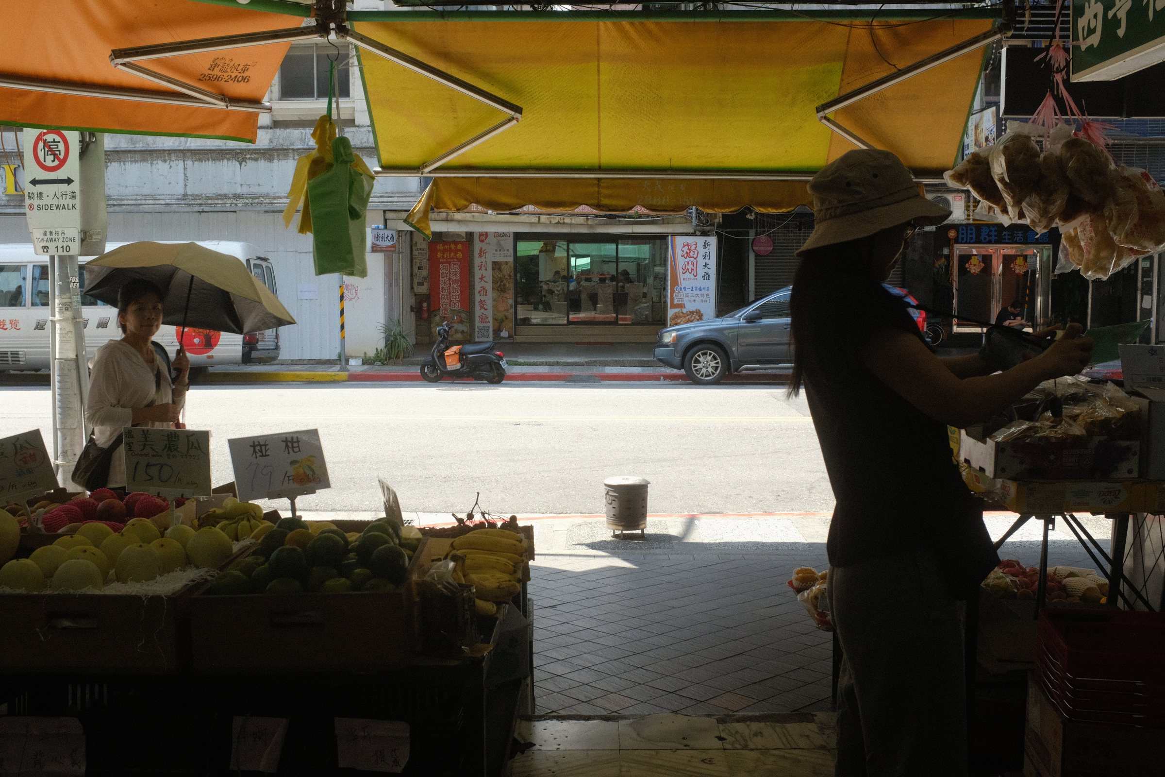 黄色い日除けの果物屋と、台に落ちる日傘の影