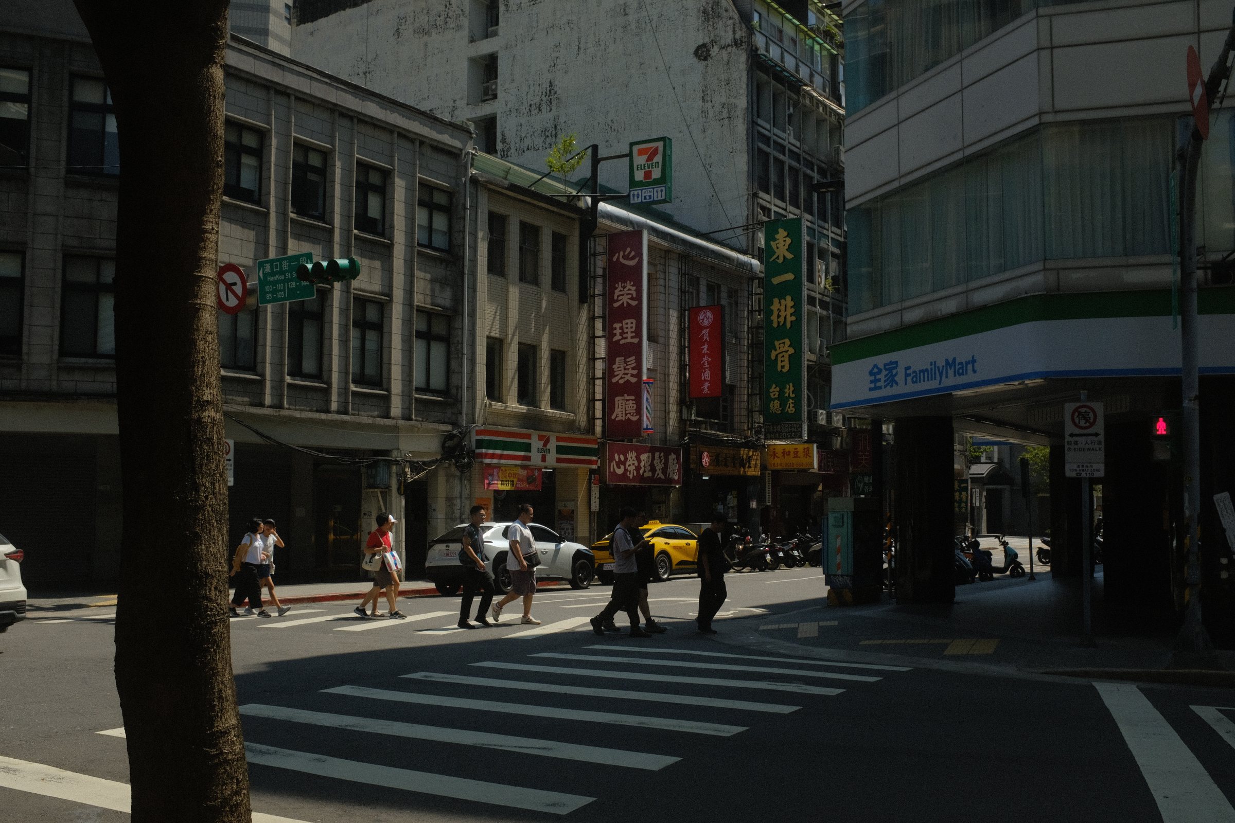 強い日差しの交差点。手前の木の幹は影になり、横断歩道を人が渡っていく