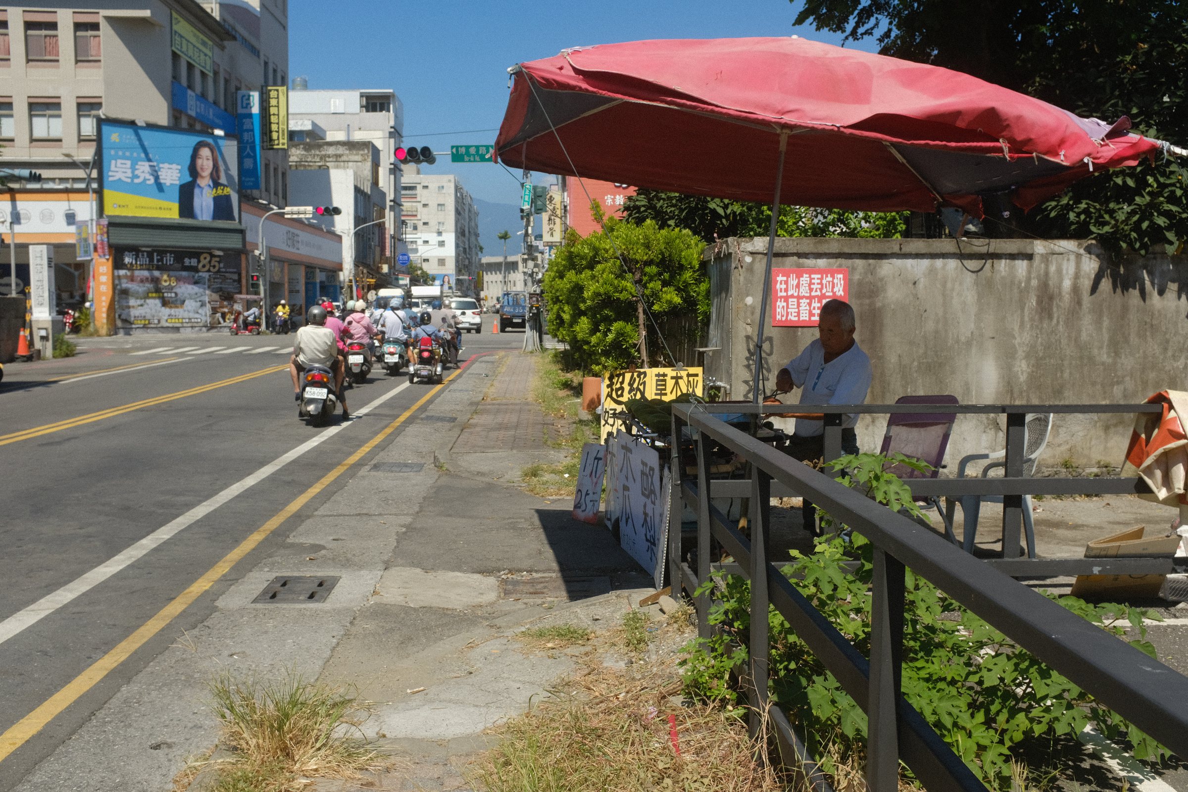 赤いパラソルの下で、道端の机に向かって座る人と、バイクの行き交う通り