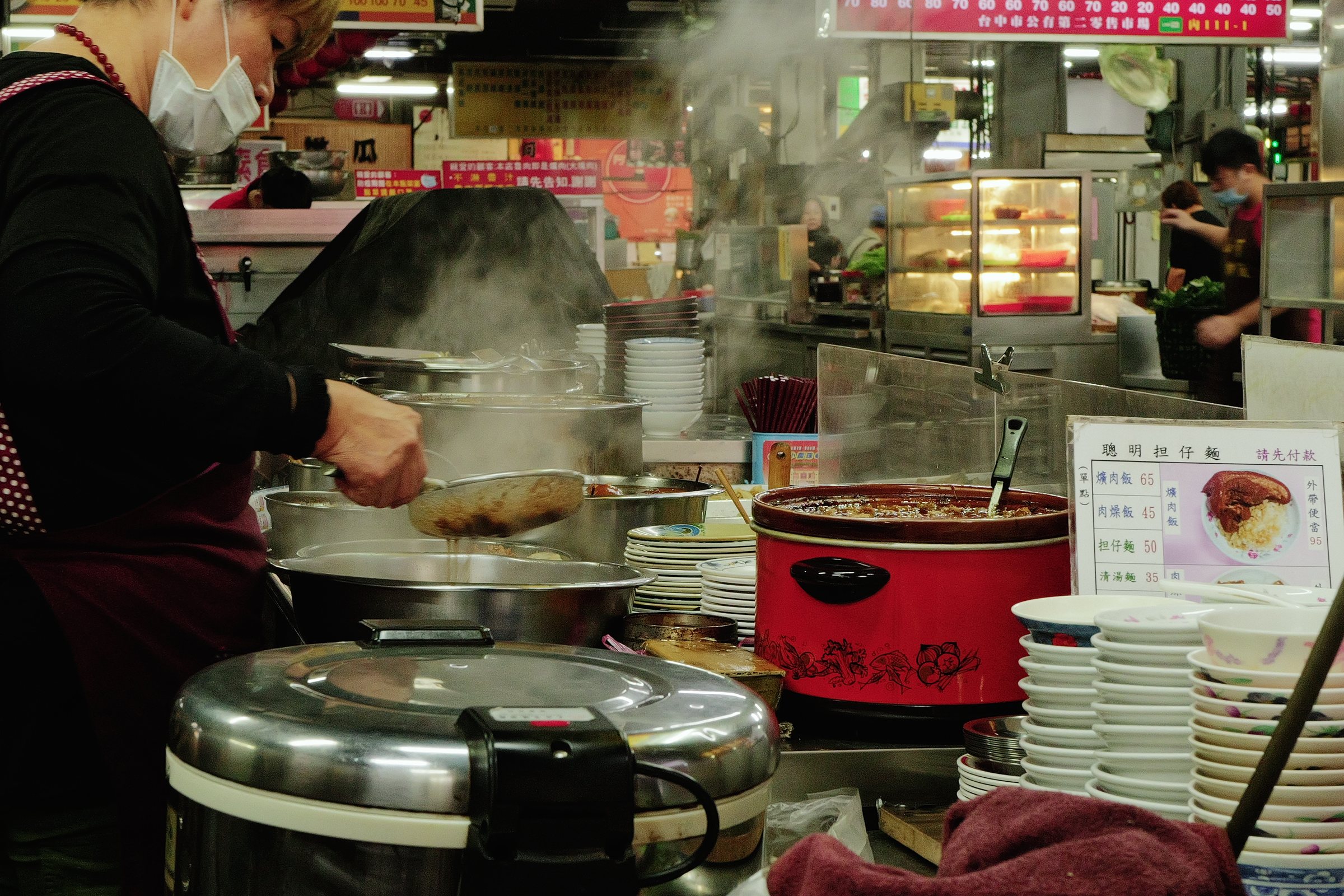 湯気の立つ食堂の厨房と、鍋の前に立つ店の人